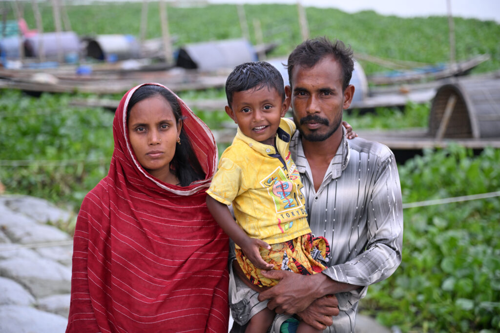 Mohammad Sumon Mridha junto a su esposa, Hena Begum, y su hijo de 3 años frente en su casa de madera y chapa, diseñada para ser desmontada y trasladada, en Louhajang Upazila, Munshiganj, Bangladesh.