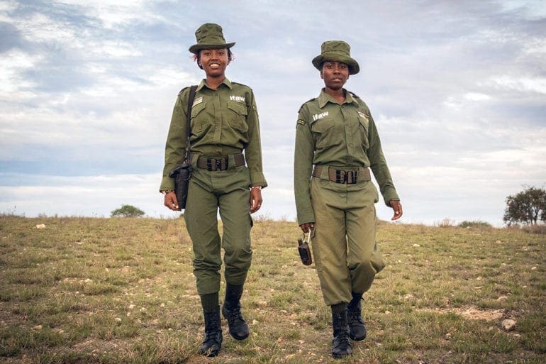 Two female Kenyan rangers dressed in green uniforms walk through the Kenyan wilderness.