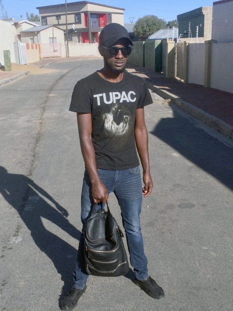 A young man wearing a black Tupac t-shirt holds a black leather backpack in one hand.