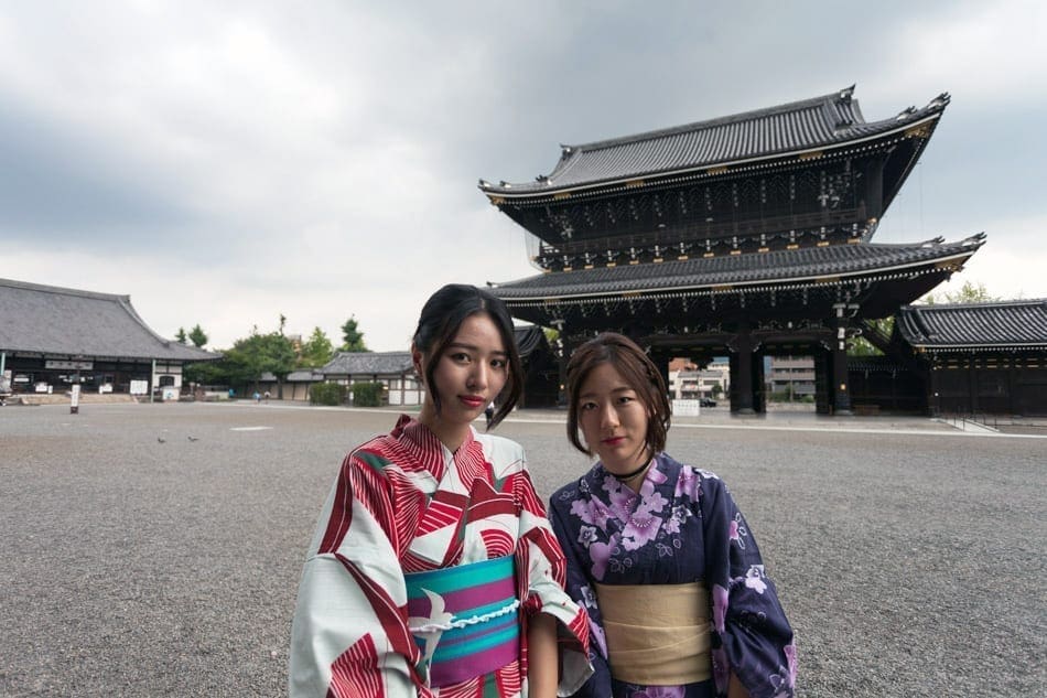 Two Japanese women in traditional clothing pose in front of a large pagoda structure.